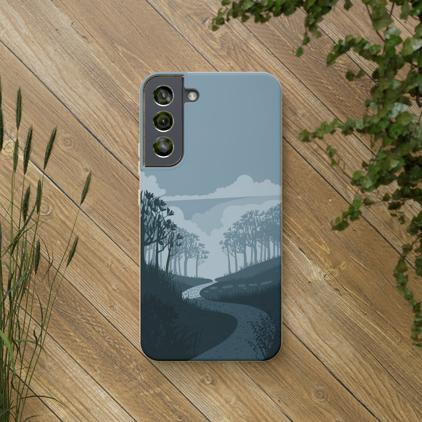 A smartphone with the Wanderlust Biodegradable Phone Case by Printify, featuring a minimalist forest path design under cloudy skies, rests on a wooden surface surrounded by green plants.
