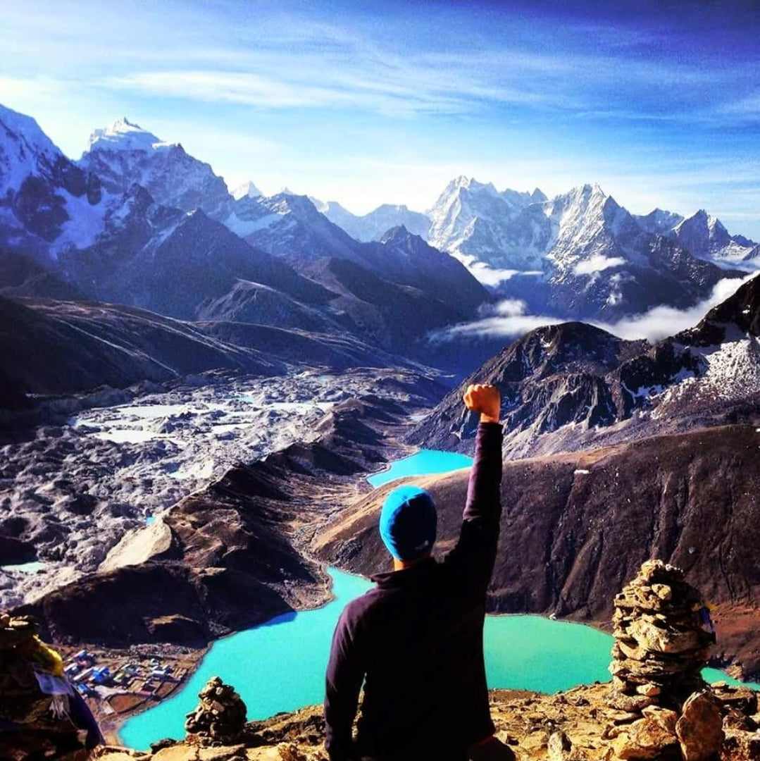 A person stands on a rocky mountain viewpoint, raising a fist triumphantly toward a panoramic landscape of turquoise lakes, rugged valleys, and snow-capped Himalayan peaks under a clear blue sky.