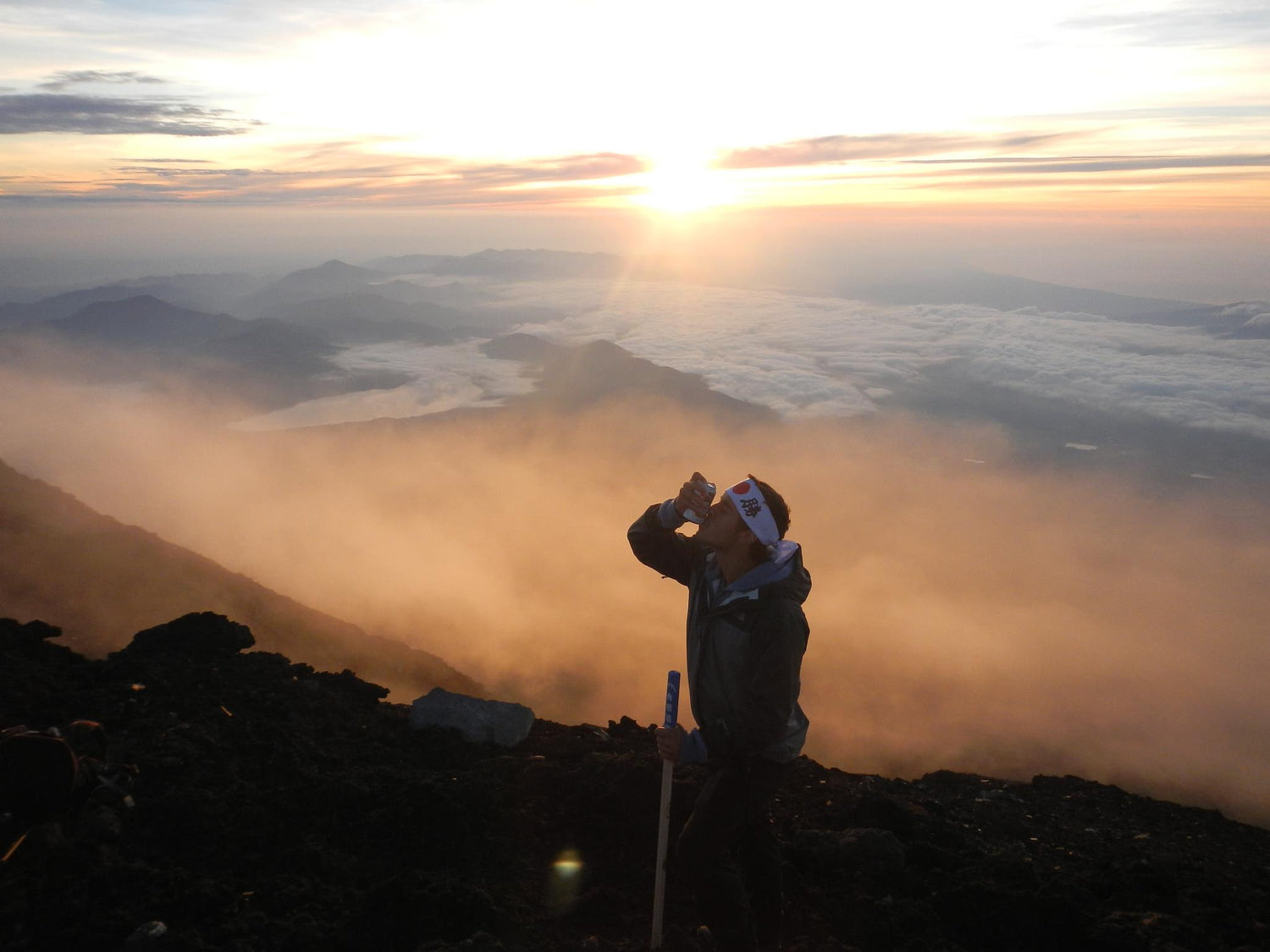 A person stands on a mountain at sunrise, holding a walking stick and drinking from a bottle. Misty clouds fill the valleys below, with warm sunlight illuminating the sky and landscape.